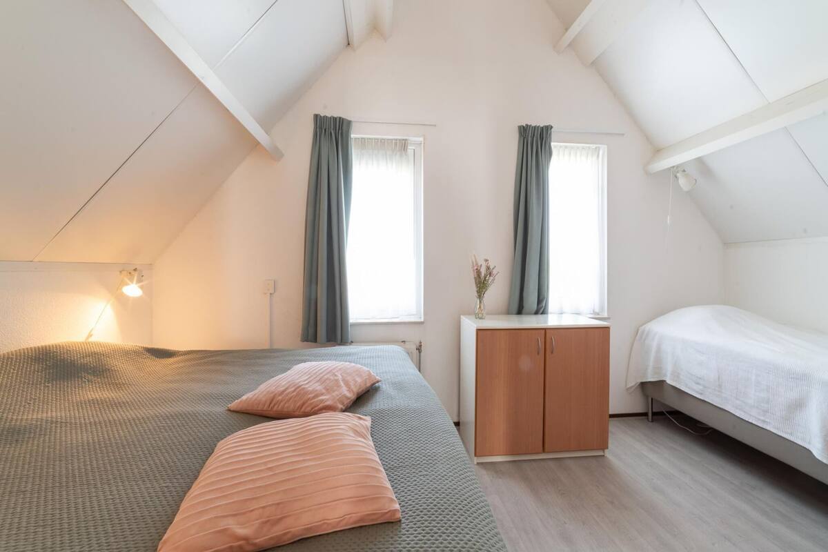 Une chambre avec un plafond en pente, un lit avec deux oreillers, une table de chevet avec un vase et une armoire.