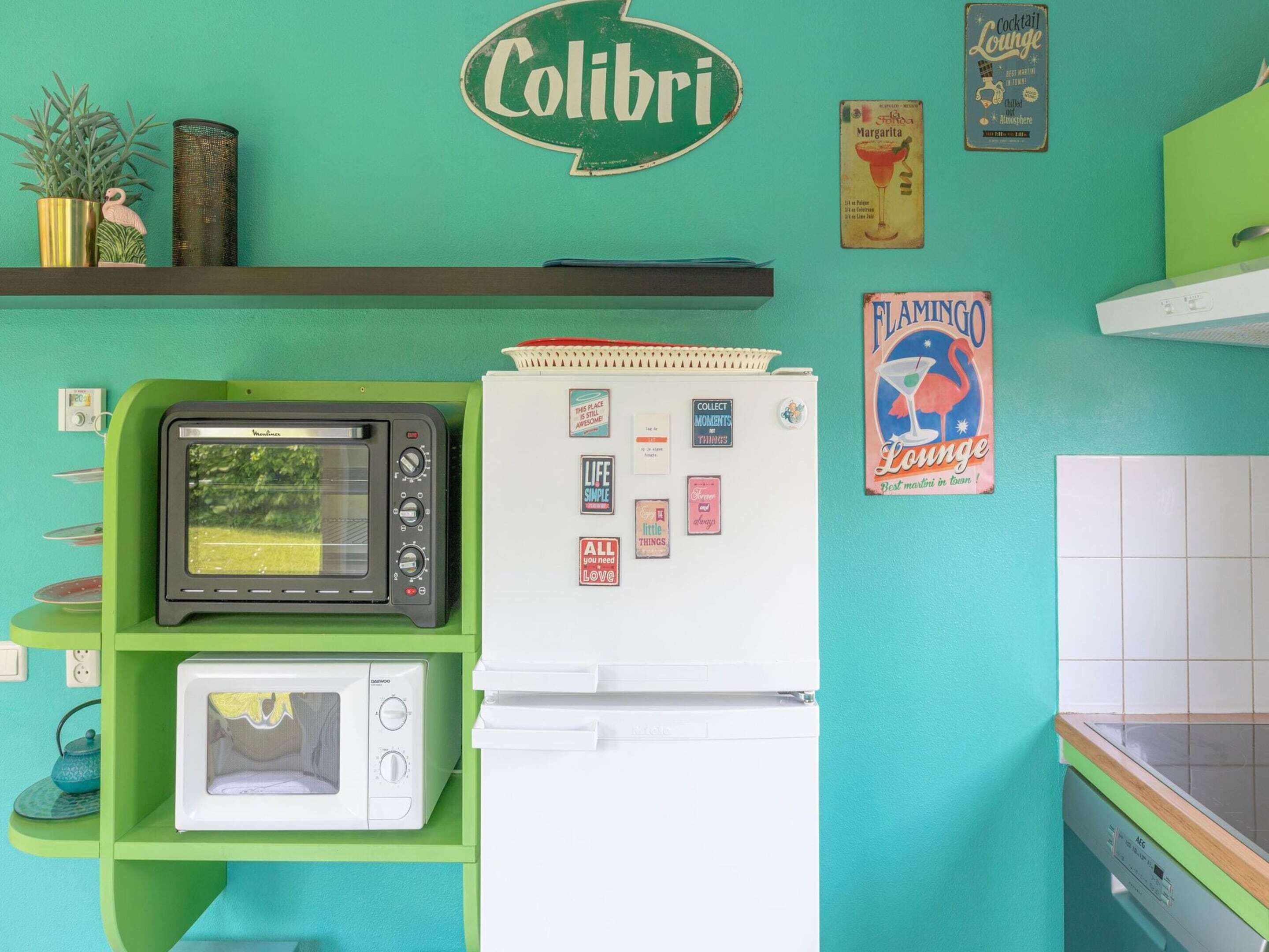 A kitchen with a green wall, a white refrigerator, a black microwave, and a white oven. There are various kitchen items and decorations on the wall.