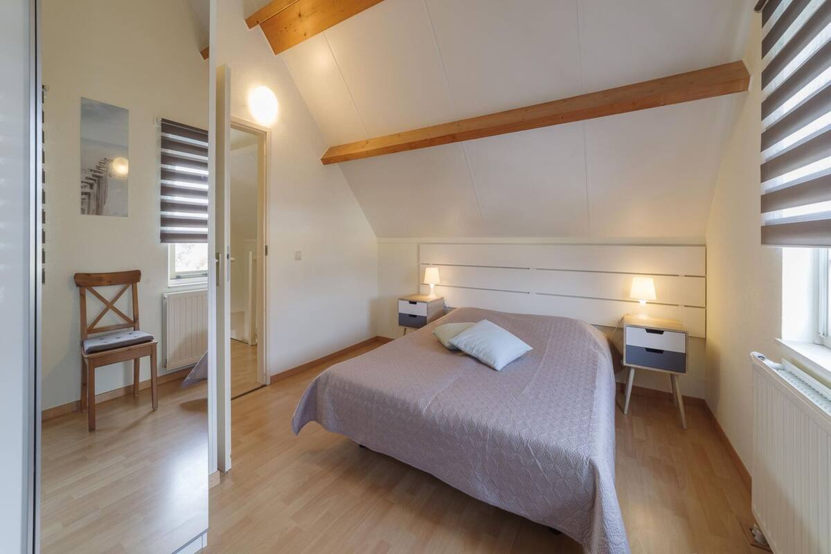 Une chambre avec un plafond en pente, des poutres en bois, un lit avec une tête de lit, des tables de chevet avec des lampes, une chaise et un radiateur.