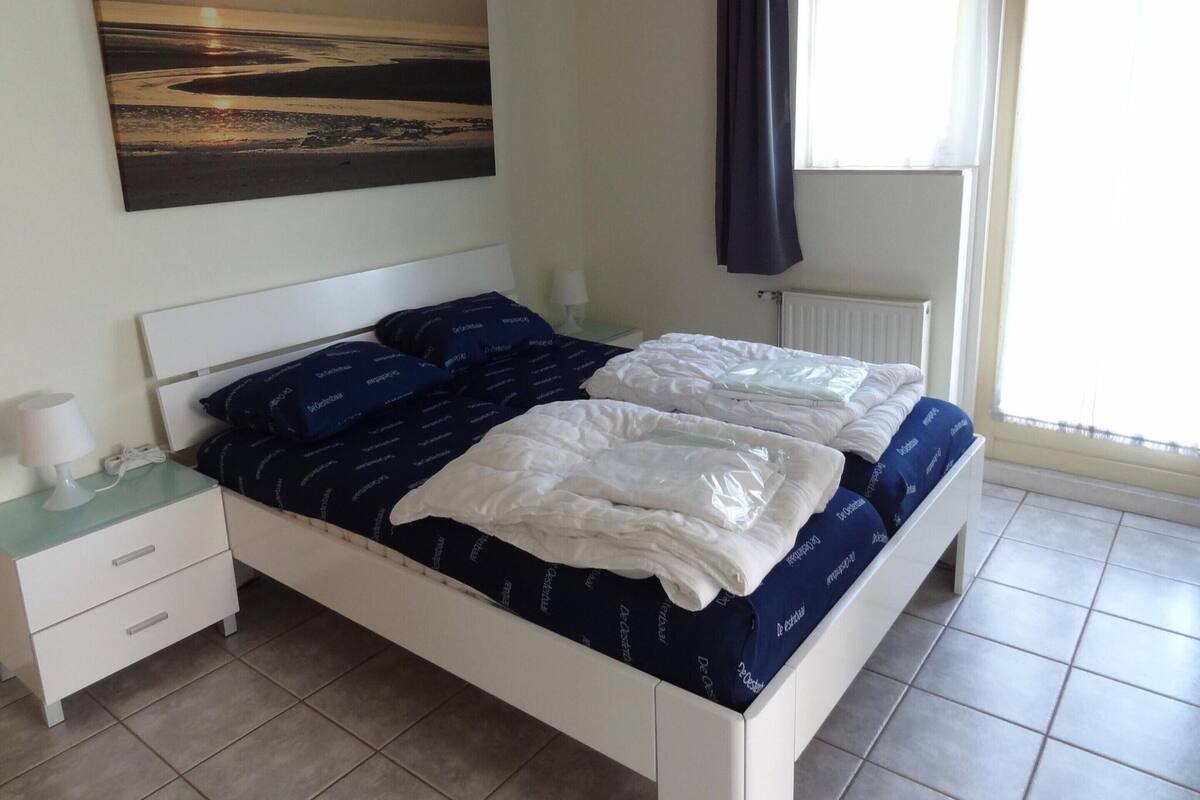 Un lit bien fait, avec une literie blanche et bleue, une table de chevet avec une lampe, et un tableau représentant une scène de plage accroché au mur.