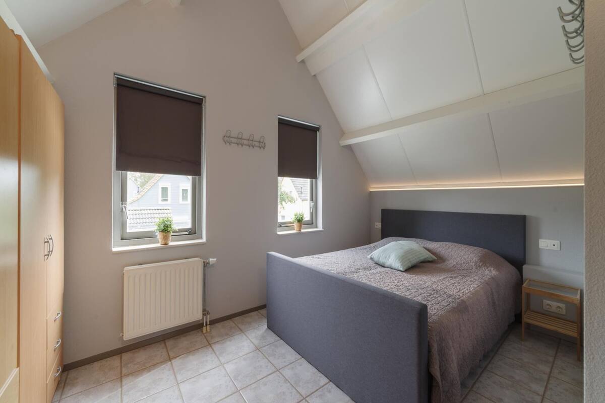 Une chambre avec un plafond en pente, deux fenêtres, un lit, une table de chevet et un radiateur.