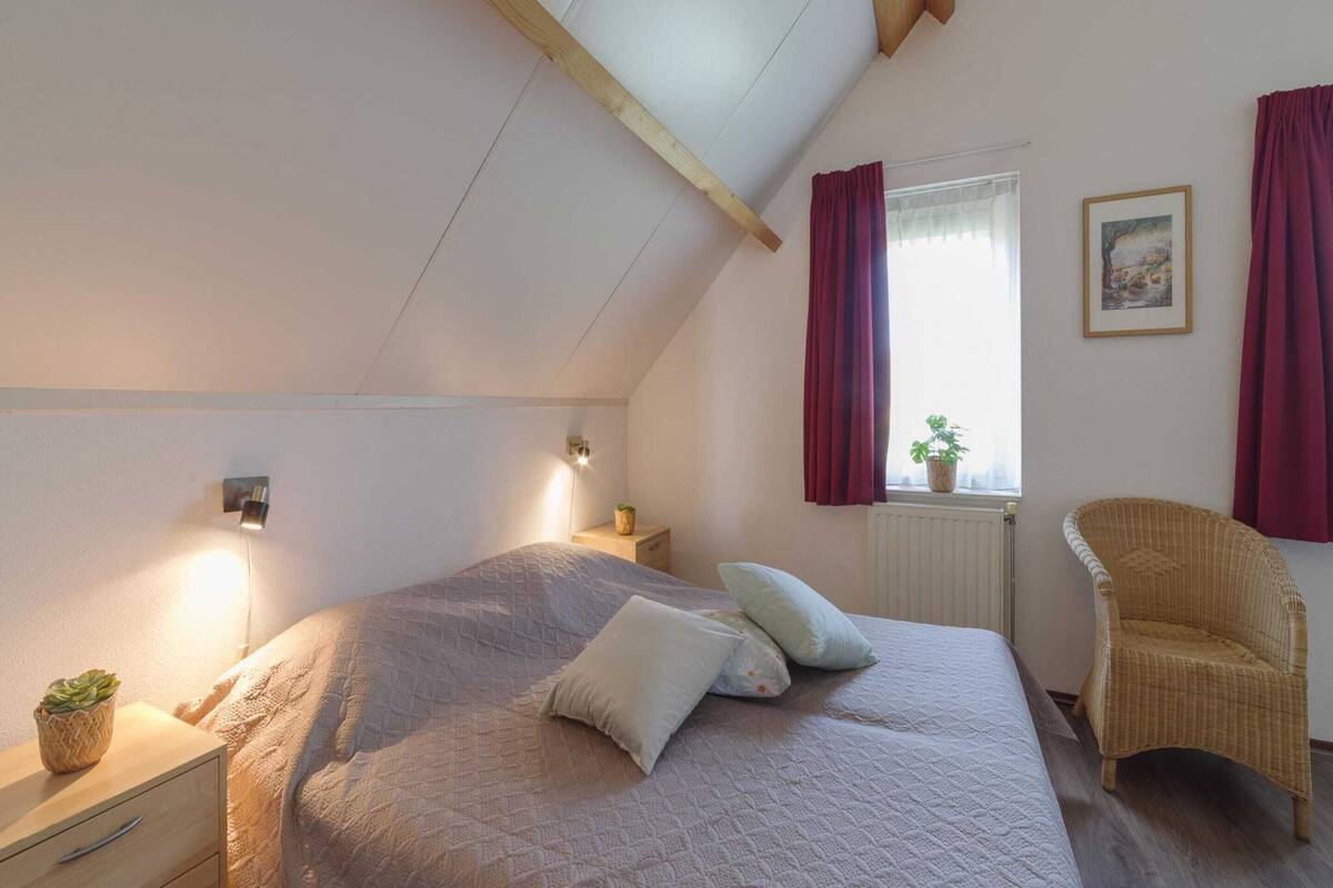 Une chambre à coucher comprenant un lit, des tables de chevet, une chaise, une fenêtre avec des rideaux et un radiateur.