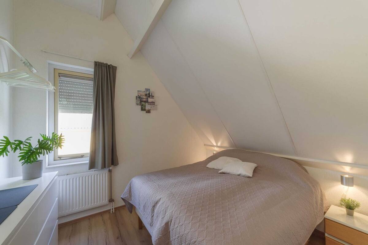 Une chambre avec un plafond en pente, un lit recouvert d’une couverture beige, une table de chevet avec une lampe, une fenêtre avec des stores et une plante en pot.