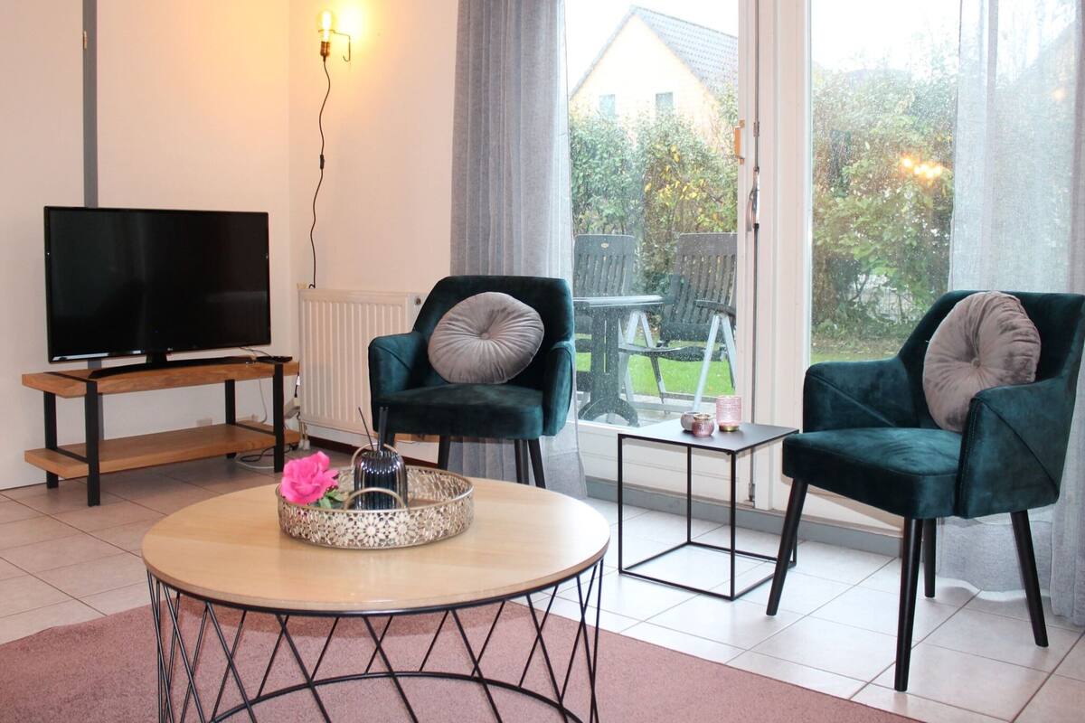 Un salon avec un téléviseur posé sur un meuble en bois, deux fauteuils verts avec des coussins, une petite table sur laquelle se trouve un plateau décoratif, et une vue sur un jardin grâce à de grandes fenêtres.