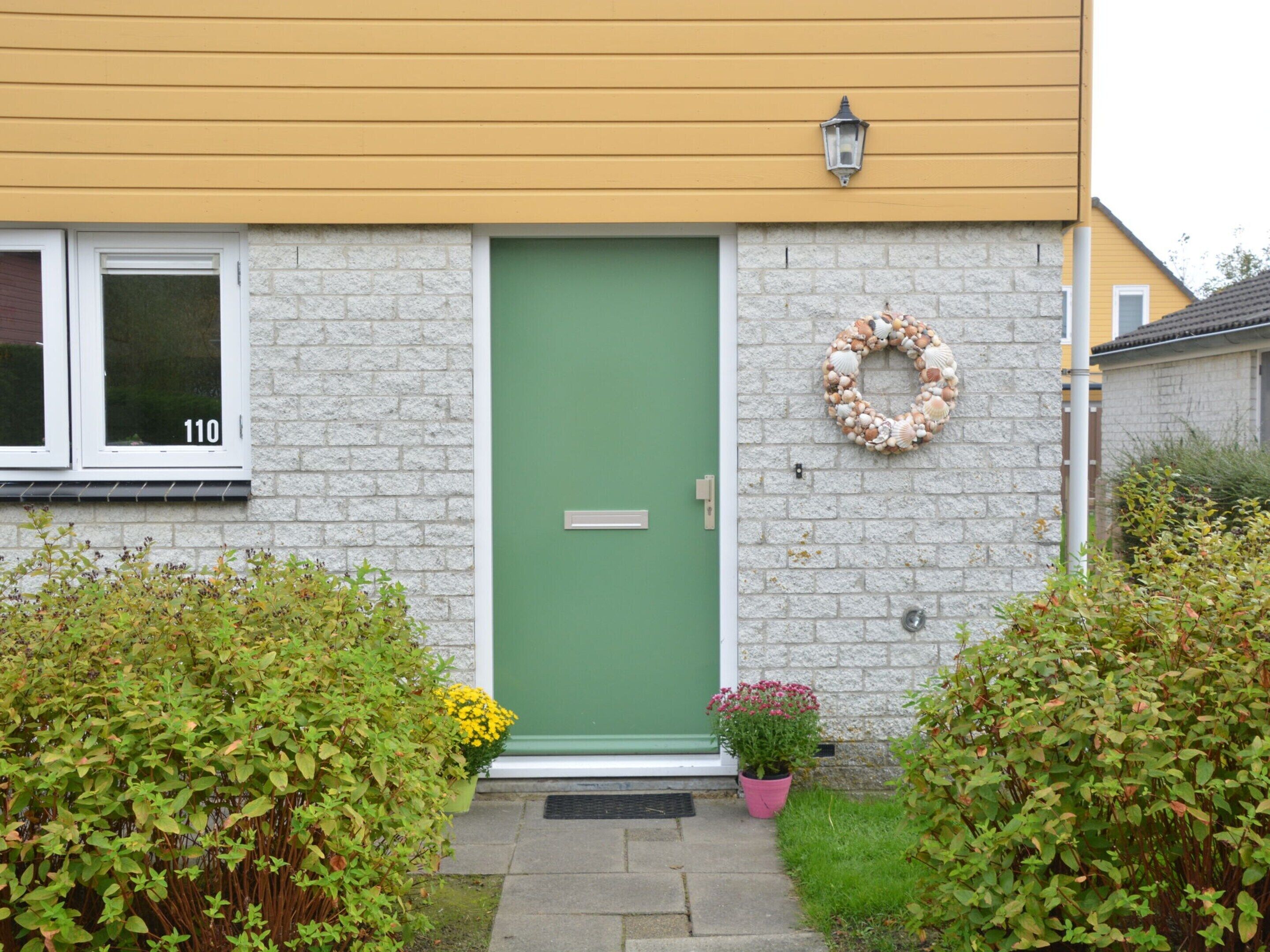 Una casa con puerta verde, el número '110' en la ventana, una corona decorativa en la pared, y un pequeño jardín con plantas y flores.