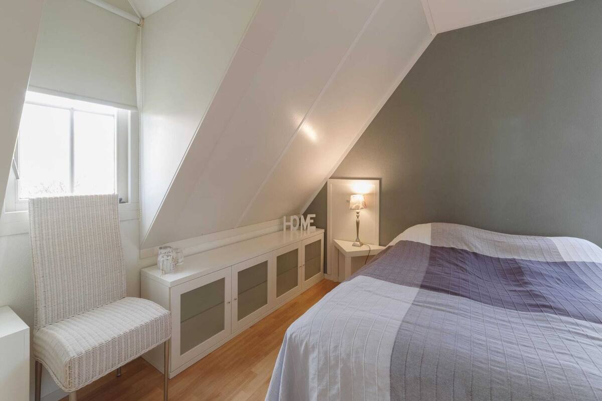 Une chambre avec un plafond en pente, un lit recouvert d’une couverture à rayures, une chaise blanche, une table de chevet avec une lampe et un meuble sur lequel on peut lire les lettres « HMF ».