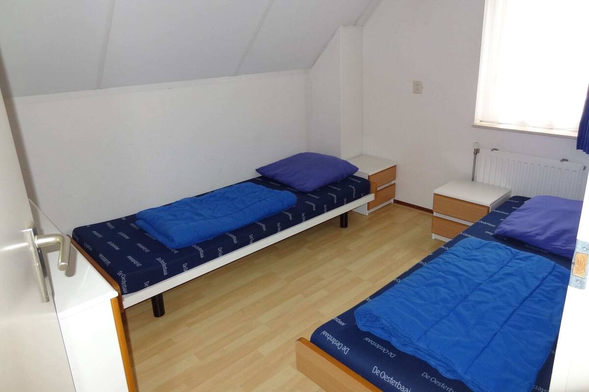 Une chambre de dortoir avec deux lits simples, chacun doté d’un matelas bleu et d’un oreiller assorti. On y trouve des tables de chevet en bois avec des tiroirs, un radiateur blanc et une fenêtre avec des rideaux.