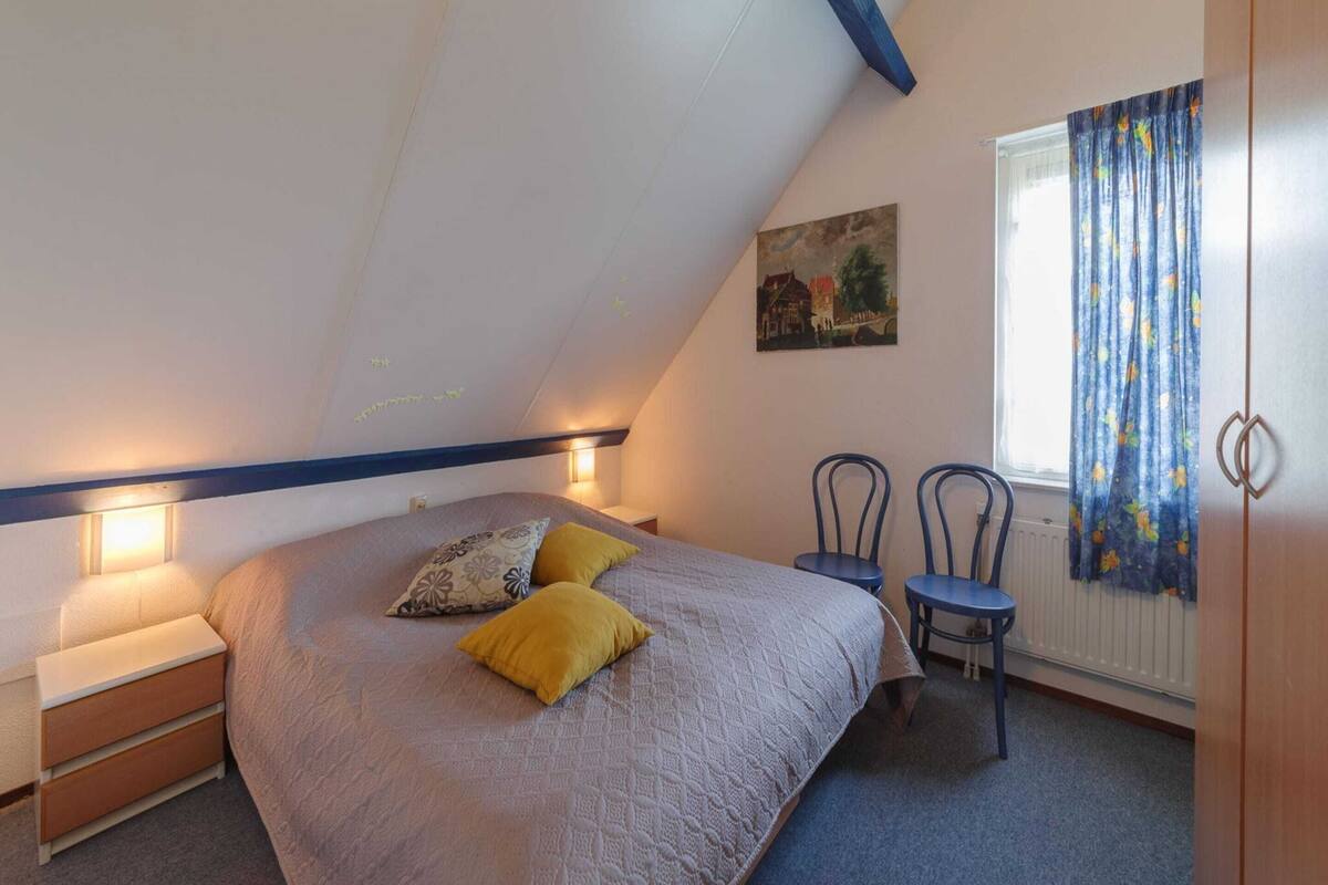 Une chambre avec un plafond en pente, un lit avec deux oreillers, une table de chevet, une chaise et une fenêtre avec des rideaux.