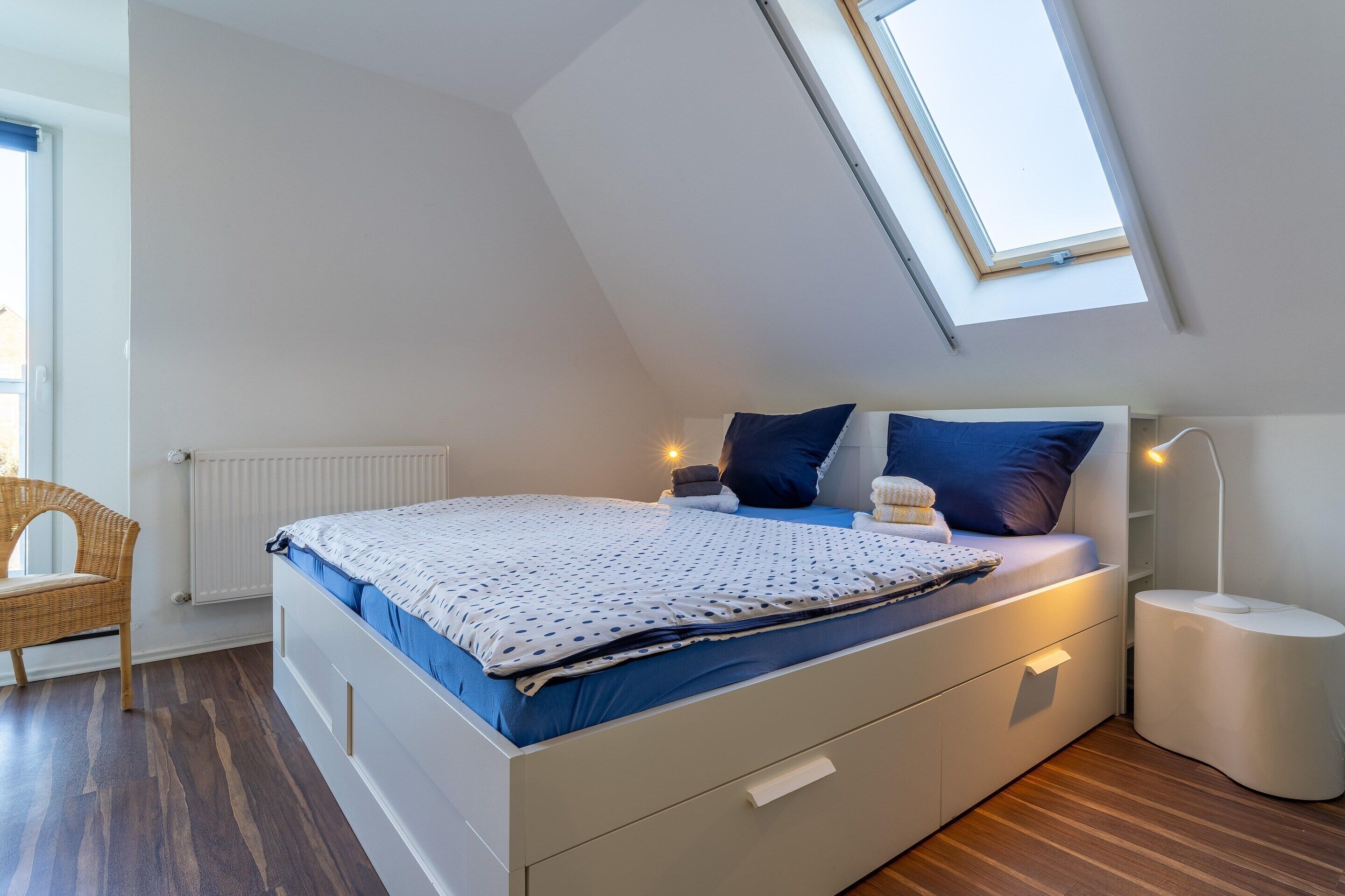 A bedroom with a sloped ceiling, a bed with a white and blue polka dot bedspread, a bedside lamp, and a window with a view of the outside.