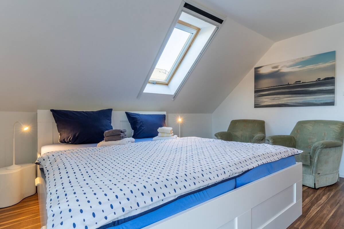 A bedroom with a skylight, a bed with a patterned duvet, a nightstand, a green armchair, and a painting on the wall.