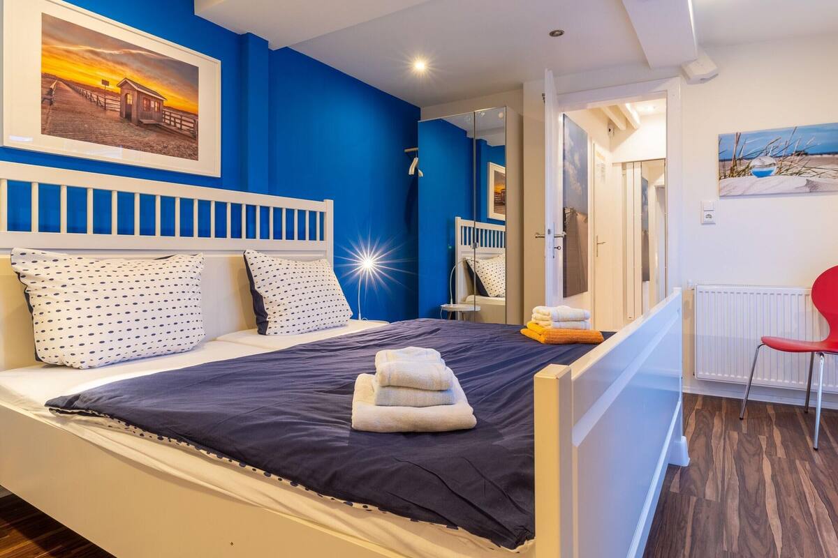 A bedroom with a bed, a nightstand, a red chair, and a painting on the wall.