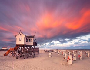 A beach house with a wooden deck and a lifeguard tower, surrounded by beach chairs and umbrellas. - Comfortable Holiday Apartment in St. Peter Ording (St. Peter-Ording)