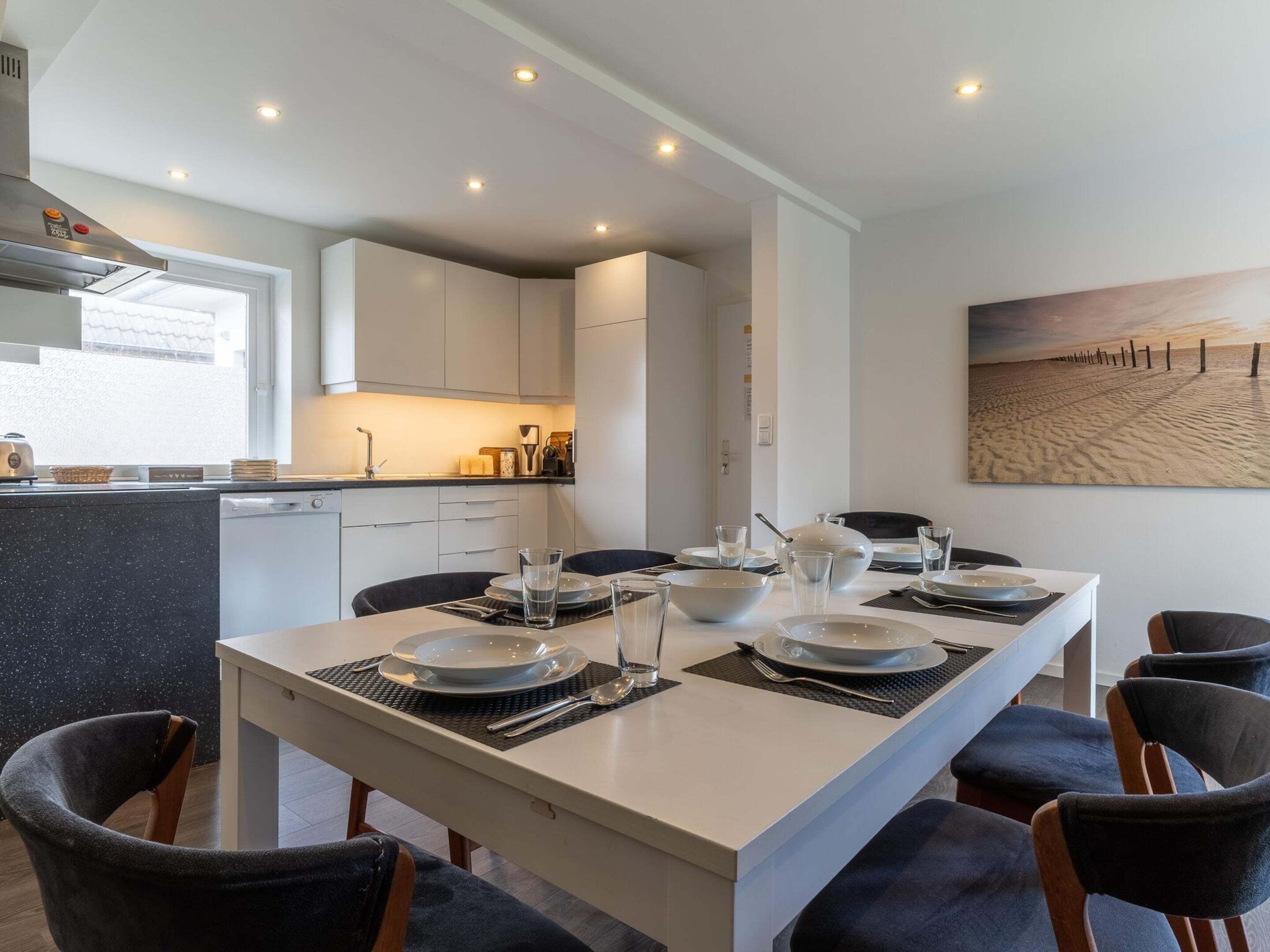 A modern kitchen with white cabinets, a dining area with a white table set for four, and a beach-themed painting on the wall.