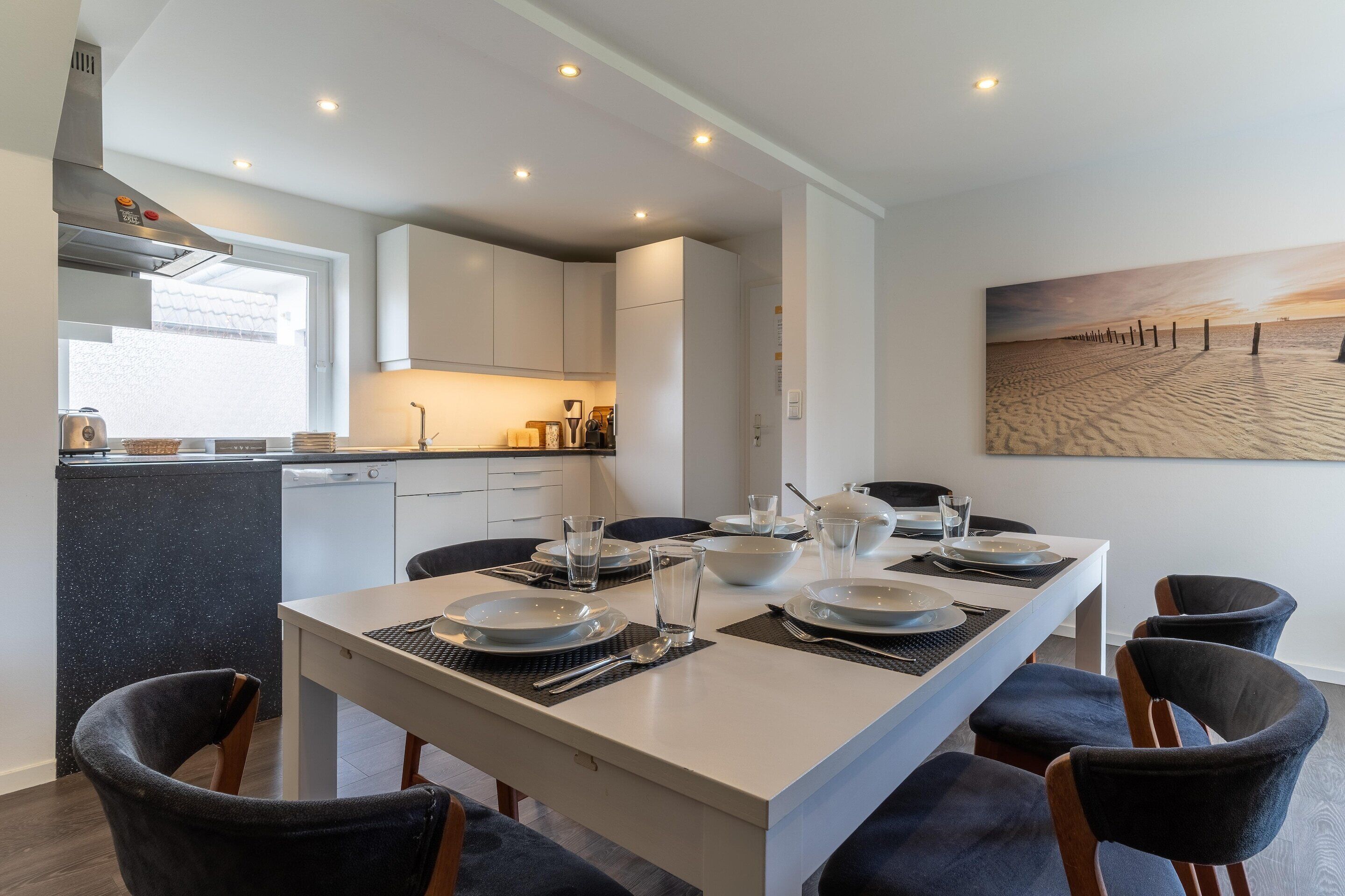 A modern kitchen with white cabinets, a dining area with a large island, and a painting of a beach scene on the wall.