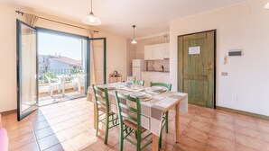 A dining area with a table set for four, green chairs, a kitchen in the background, and a sliding glass door leading to an outdoor patio. - Residence I Mirti Bianchi, Santa Teresa Gallura (Santa Teresa di Gallura)