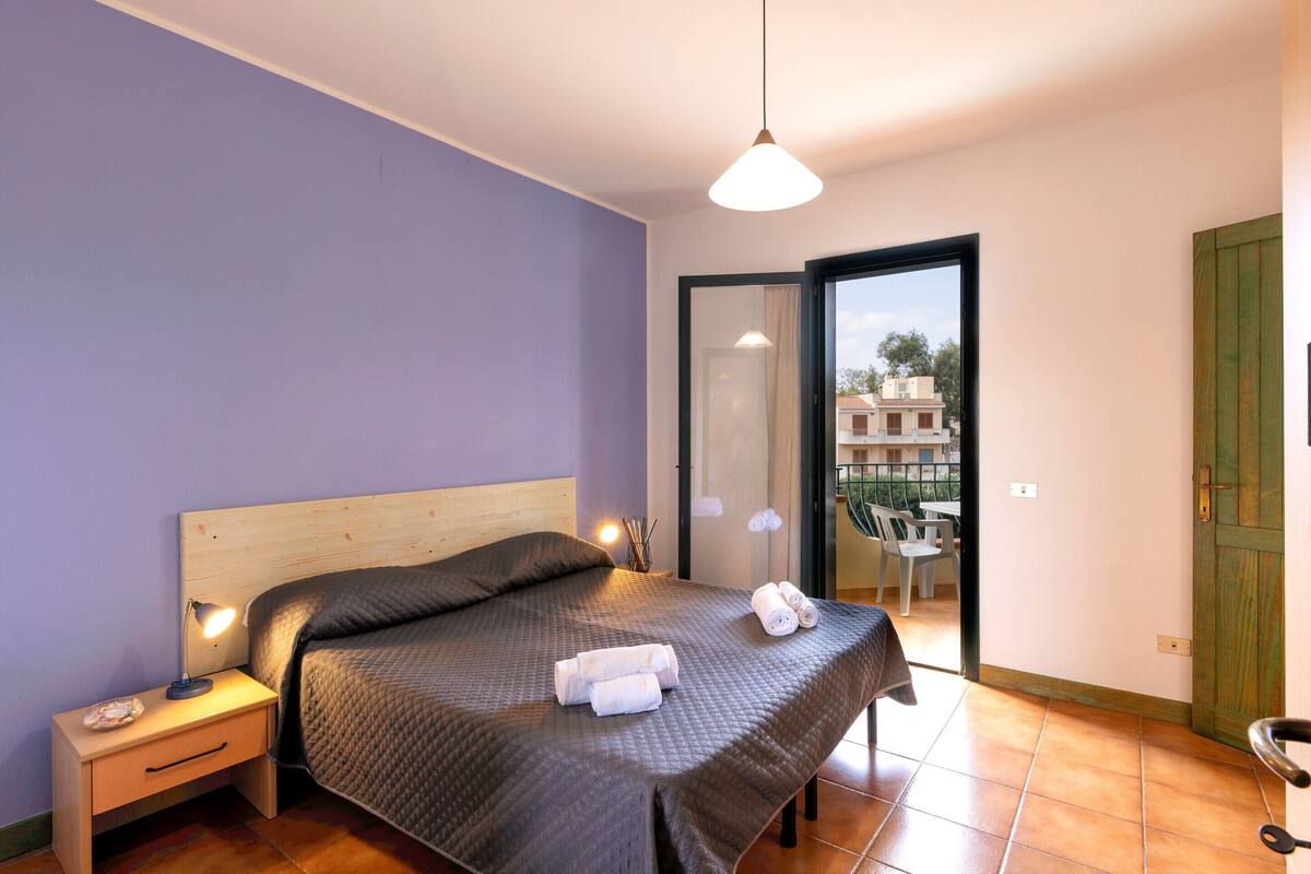 Ein Schlafzimmer mit Bett, Nachttisch, Lampe und Blick ins Freie.