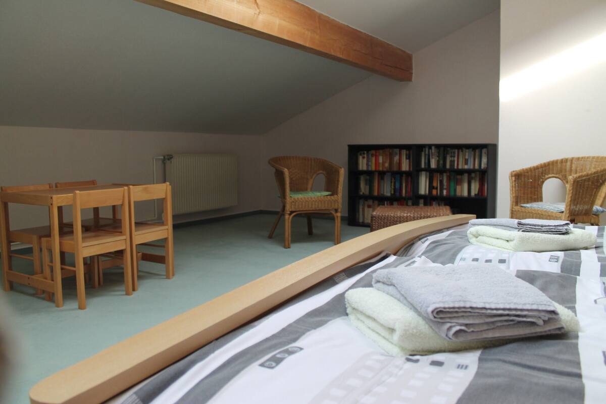 Une pièce comprenant un lit, deux chaises en bois, un fauteuil en osier et une bibliothèque.