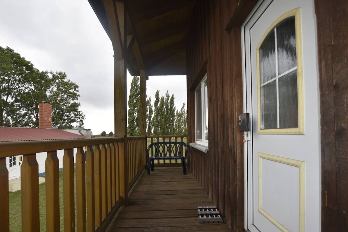 A wooden porch with a bench, a door with a number, and a view of trees.