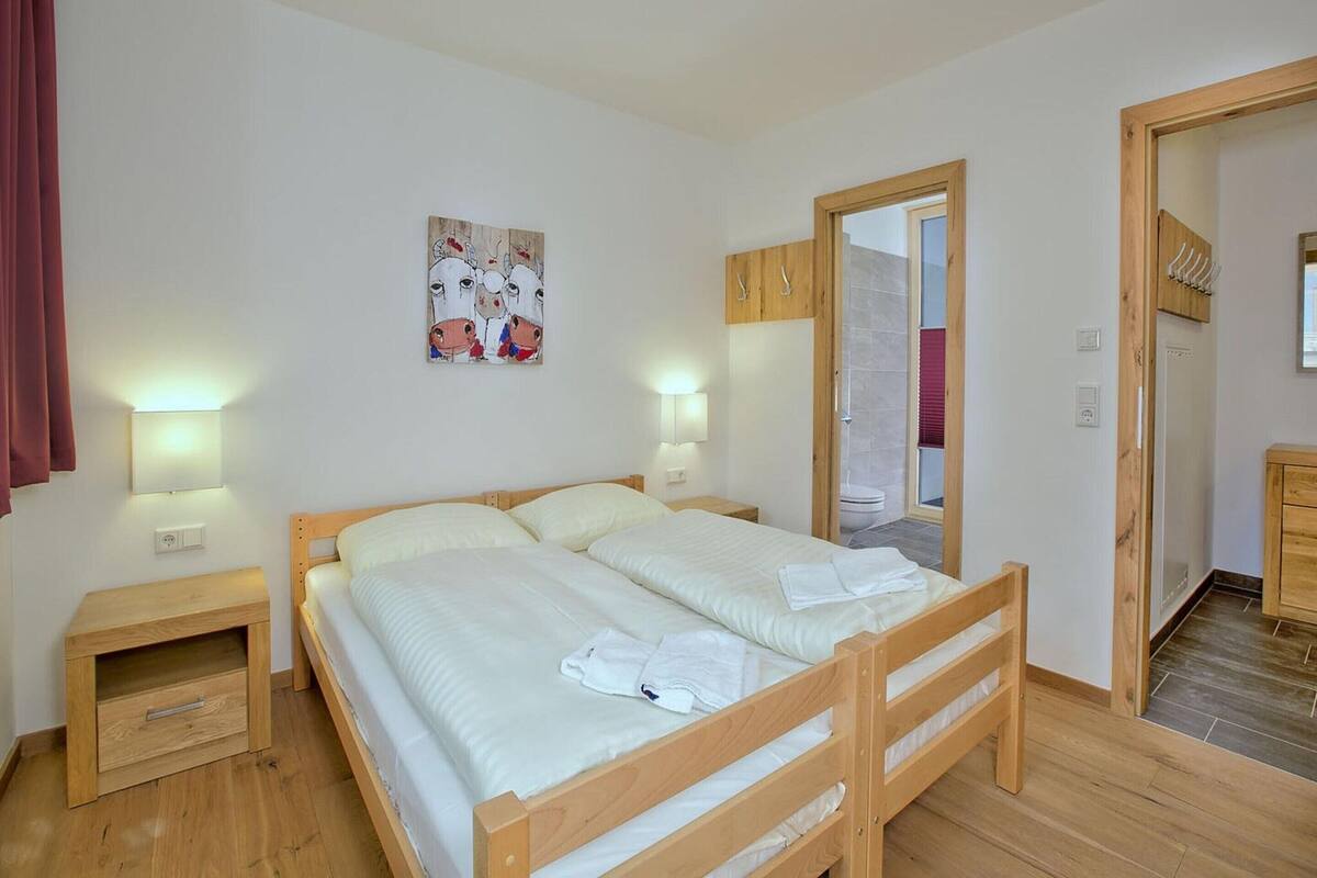A double bed with white linens, two bedside tables, a wooden headboard, a wall-mounted painting, a wooden wardrobe, and a bathroom with a shower and toilet visible through an open door.