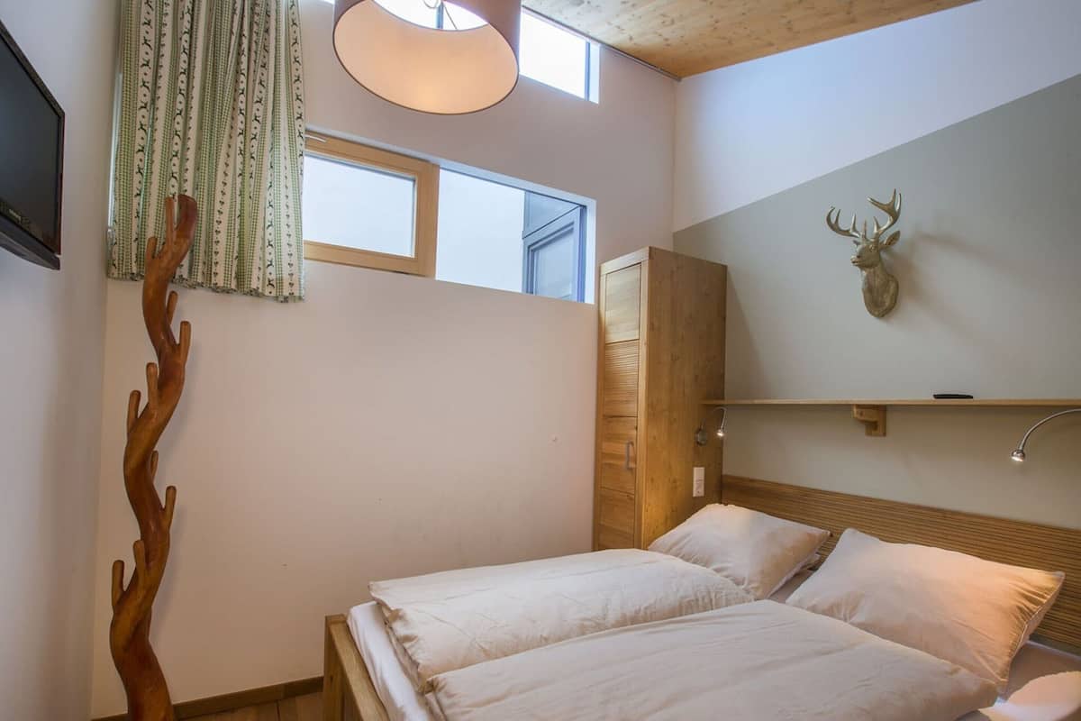A bedroom with a wooden bed, a nightstand, a wall-mounted lamp, a window, and a deer head decoration.