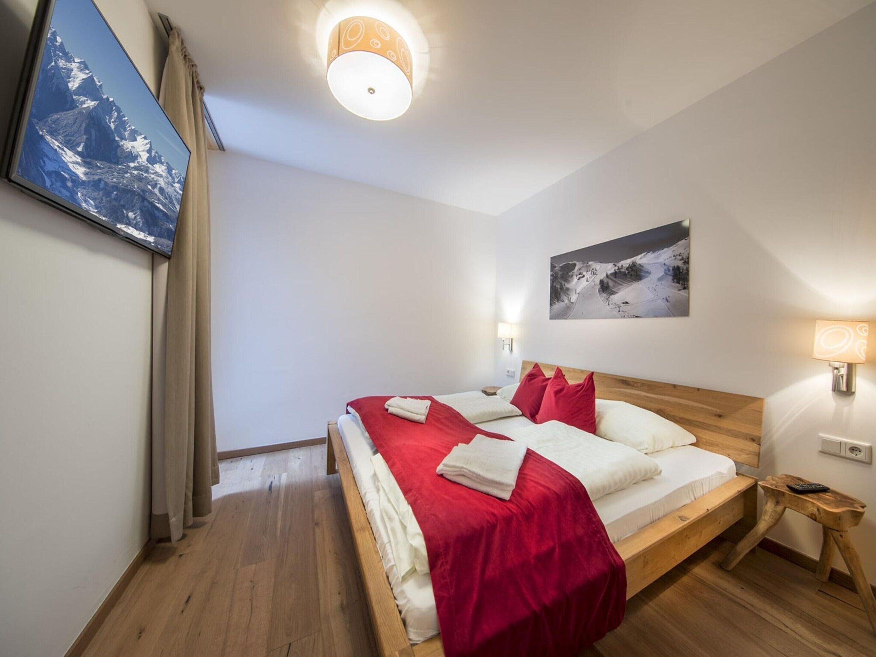A bedroom with a wooden bed, red and white bedding, a mounted TV, and a painting of a mountain.
