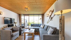 A modern living room with a wooden ceiling, a flat-screen TV, grey sofas, a coffee table, and a large window with a view of the outdoors. - Modern Apartment With Balcony and Access to Sauna (Neukirchen am Grossvenediger)
