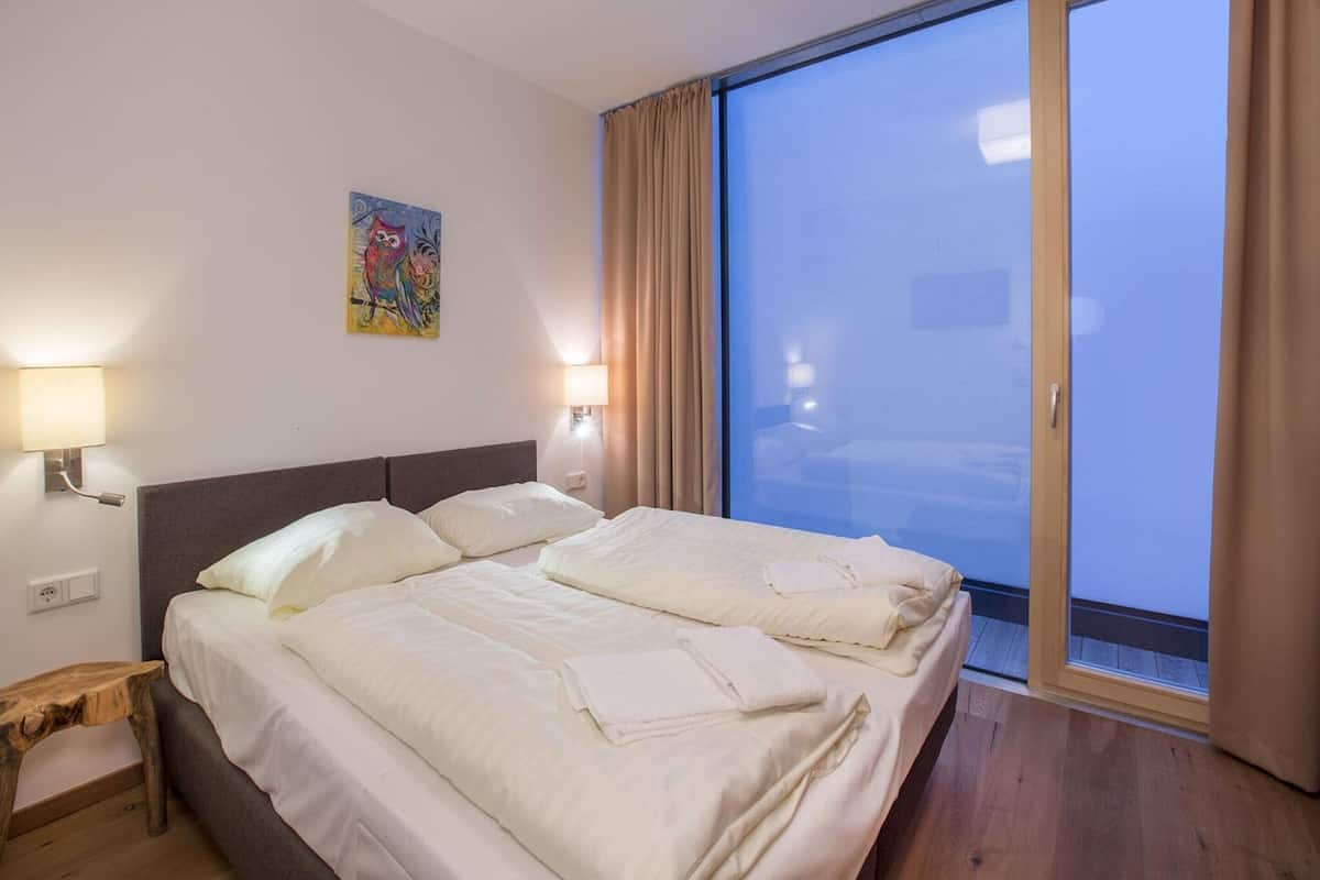 A hotel room with a large bed, two bedside lamps, a small wooden stool, a colorful painting, and a sliding glass door to a balcony.