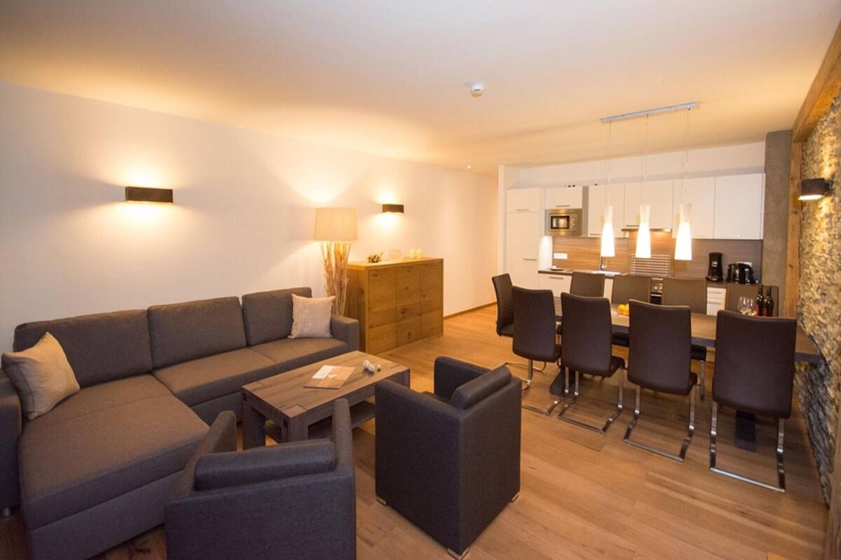 A modern living room with a sofa, coffee table, and chairs, a wooden dining table with chairs, a kitchen area with cabinets and appliances, and a stone accent wall.