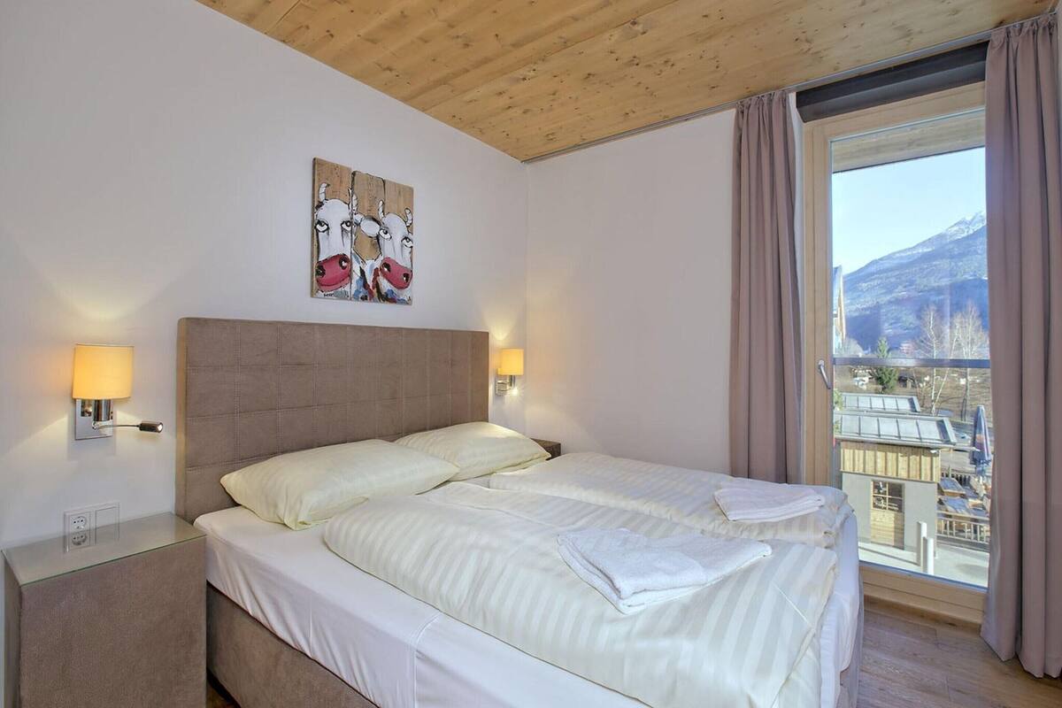 A hotel room with a bed, bedside lamps, a painting, and a view of a city and mountains.