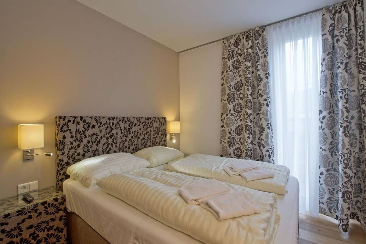 A hotel room with a double bed, two bedside lamps, a nightstand with a lamp, floral-patterned headboard, and a window with sheer curtains.