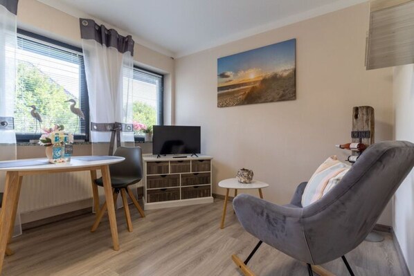 A modern living room with a grey armchair, a small round table, a TV on a wooden stand, and a bookshelf.