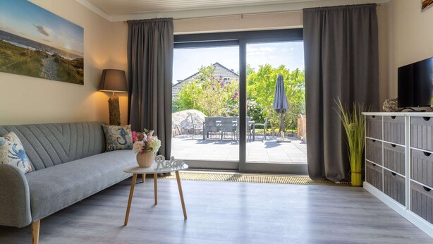 A living room with a grey sofa, a small table with a vase, a TV on a white cabinet, and a view of a garden through large sliding doors. - Comfortable Holiday Apartment in St. Peter Ording (St. Peter-Ording)