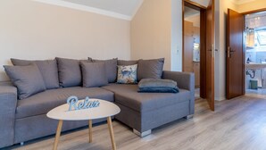 A modern living room with a grey sofa, a small white table with a 'Relax' sign, wooden flooring, and a wooden door leading to a bathroom. - Comfortable Holiday Apartment in St. Peter Ording (St. Peter-Ording)