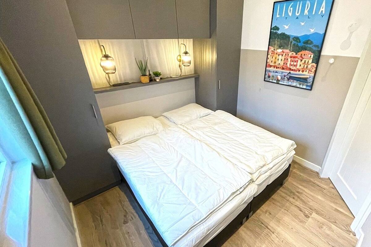 Une chambre à coucher avec un lit, une tête de lit agrémentée de lampes murales, une affiche représentant la Ligurie, en Italie, et un plancher en bois.