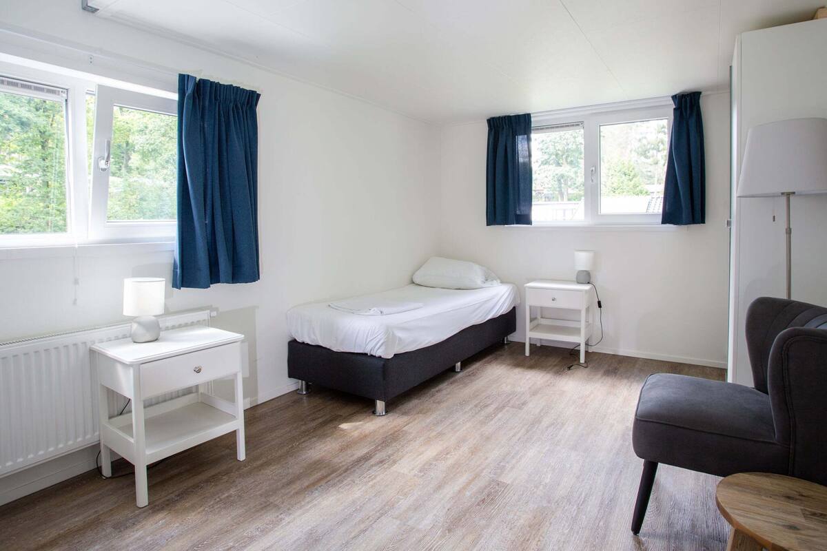 Une chambre individuelle comprenant un lit, une table de chevet, une chaise et une fenêtre avec des rideaux.