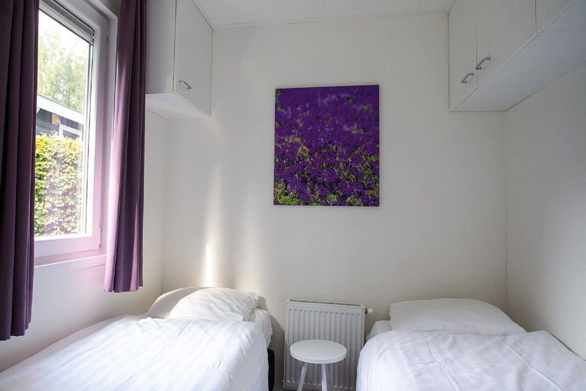 Une chambre d’hôtel avec deux lits simples, un radiateur, une petite table blanche et une fenêtre avec des rideaux.