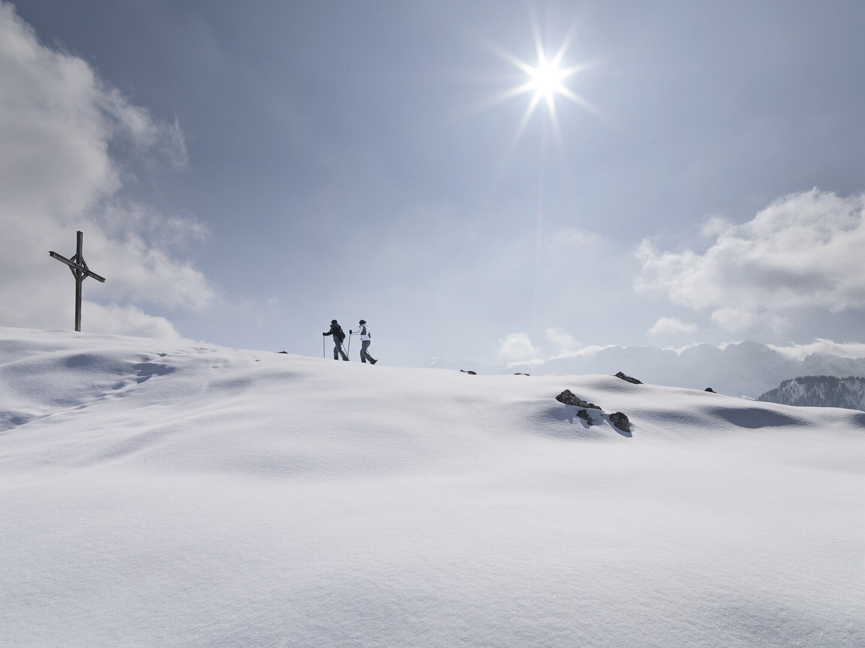 A snowy landscape with a wooden cross and two people hiking.