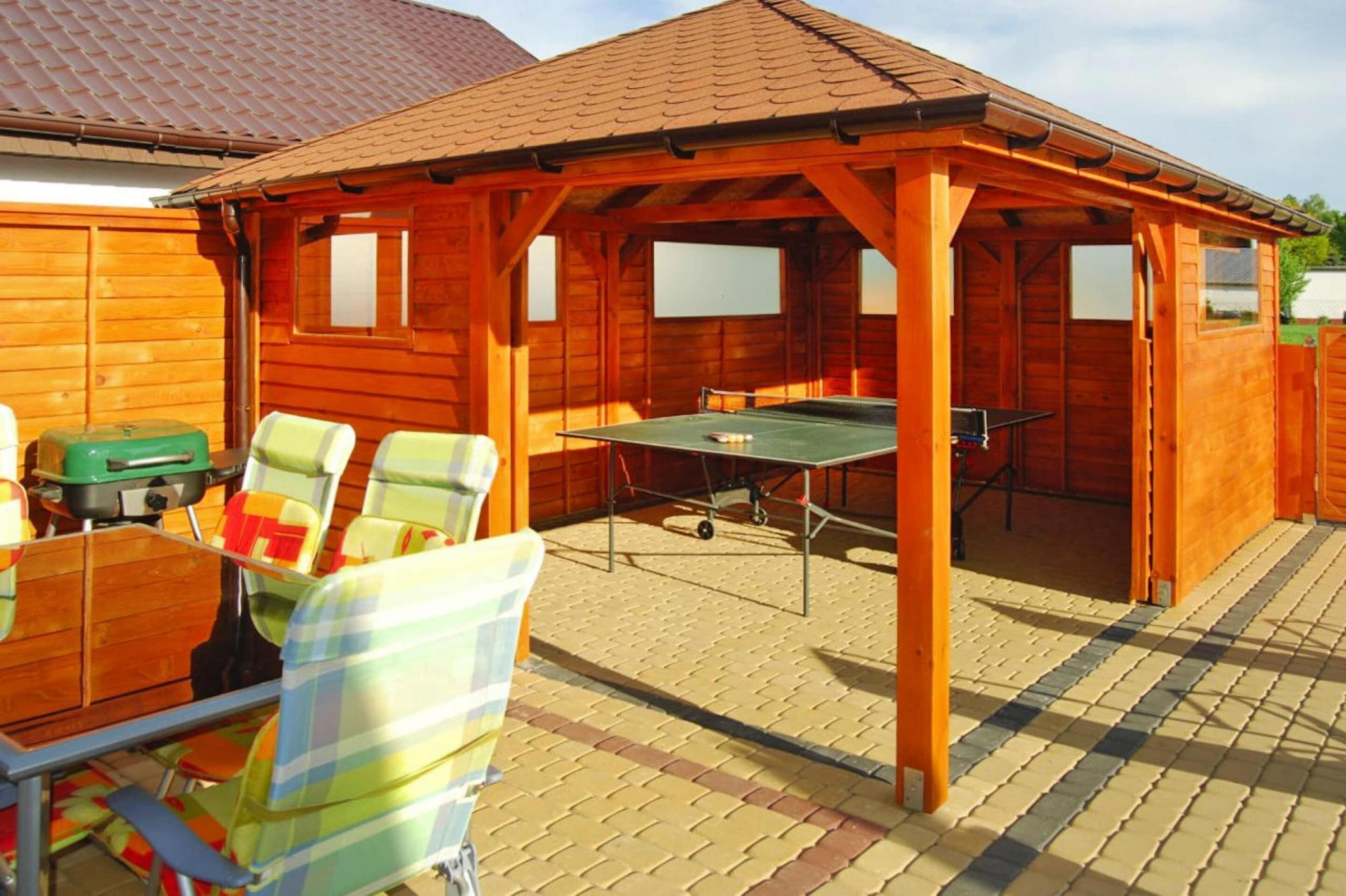 A wooden outdoor patio with a ping pong table, chairs, and a table with a green tablecloth.