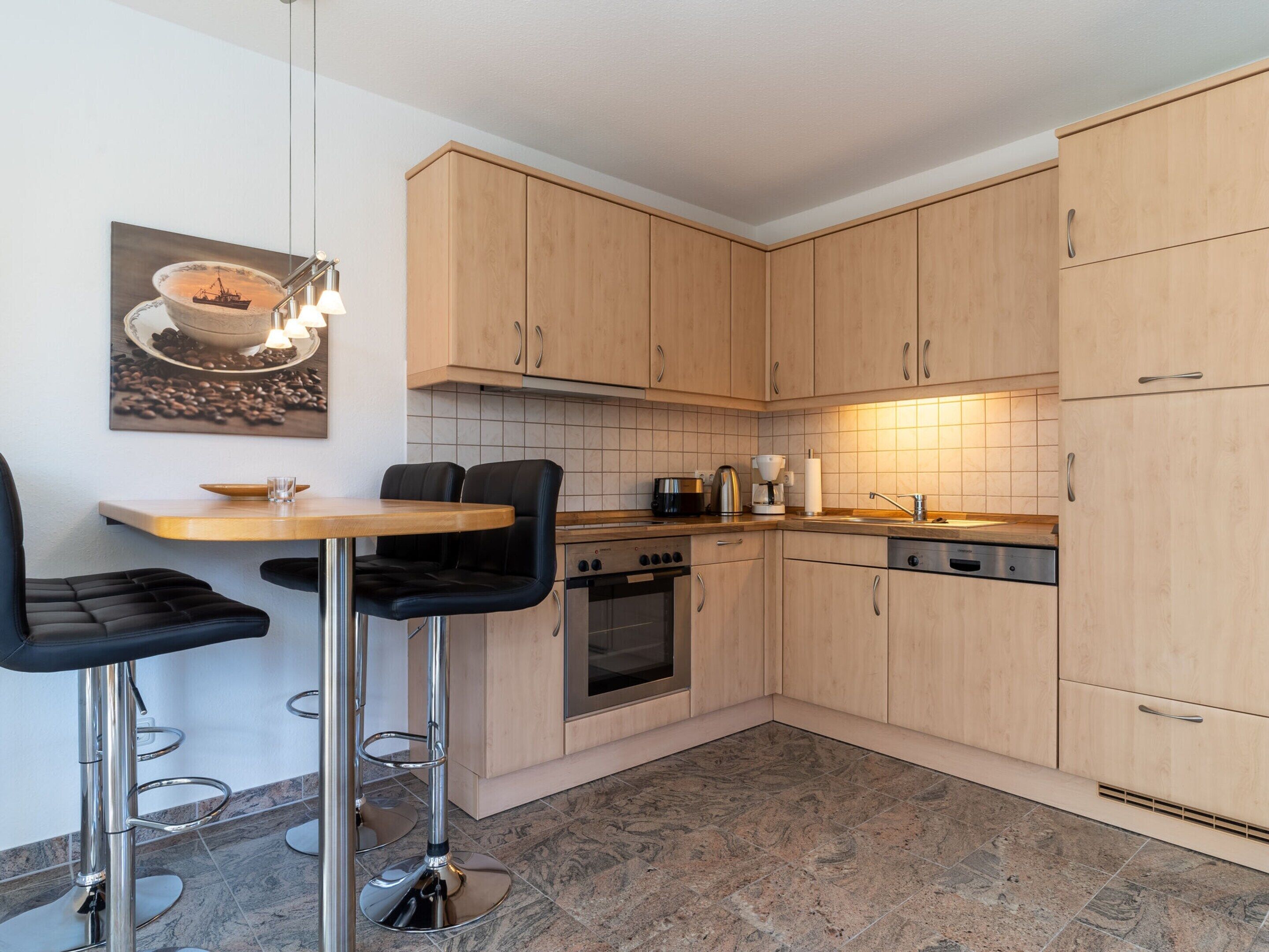 A modern kitchen with wooden cabinets, a dining table, two bar stools, a coffee maker, and a tiled backsplash.