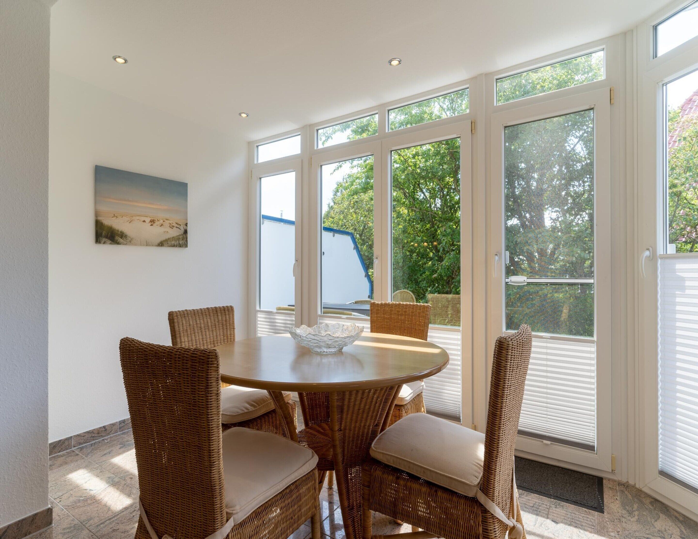A dining area with a round table and wicker chairs, large windows with a view of greenery, and a painting on the wall.