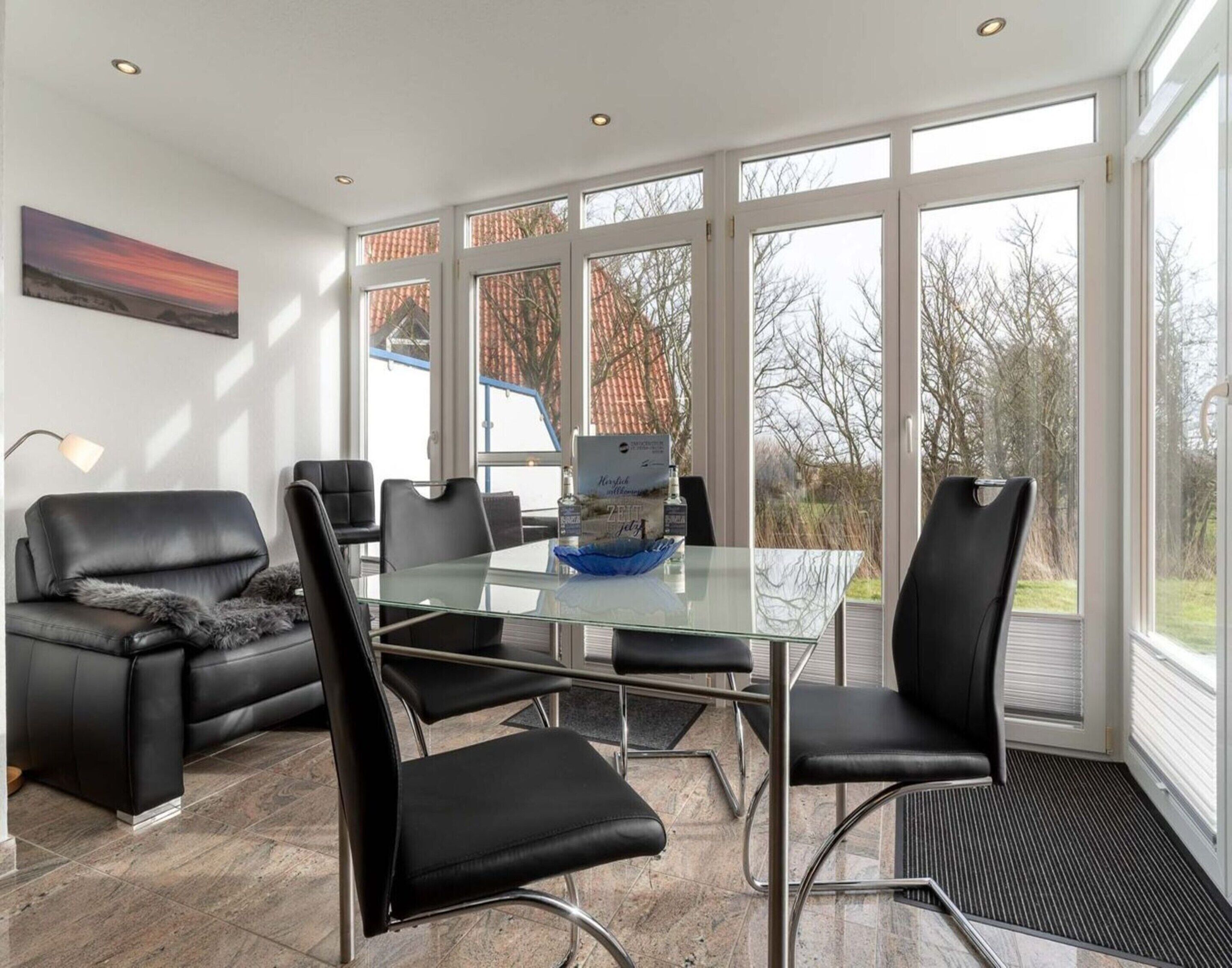 A modern living room with a glass table, black leather chairs, a black leather sofa, and a large window with a view of trees and a building.