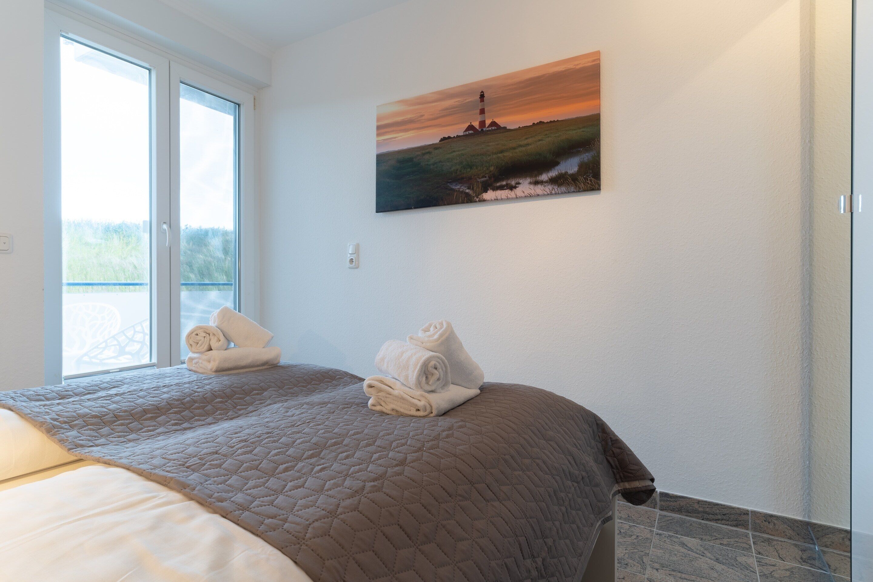 A hotel room with a large bed, a painting of a lighthouse, and a view through a sliding door.