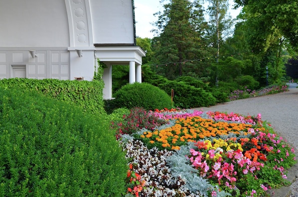 Un bâtiment blanc doté d’un porche abrité, entouré d’une végétation luxuriante et agrémenté d’un parterre de fleurs aux couleurs vives.