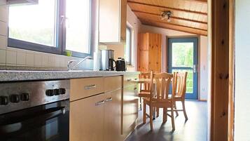 A modern kitchen with a built-in oven, stainless steel appliances, and wooden cabinets. There is a dining area with a table and chairs, and a window providing natural light.