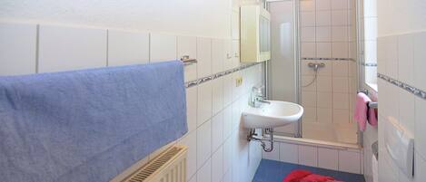 Un baño con un lavamanos blanco, una mampara de ducha de vidrio, una toalla azul y un tapete de baño rojo.