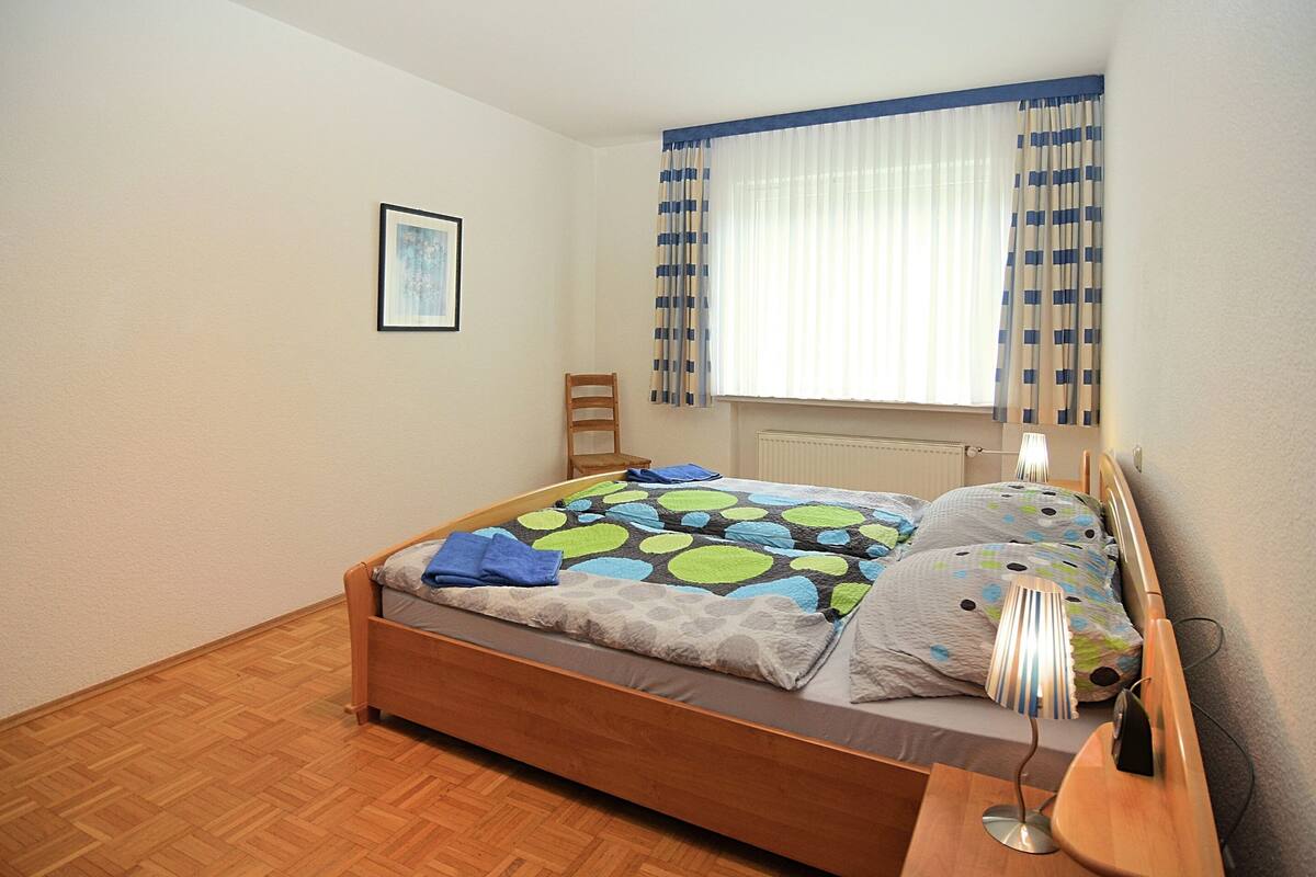 Ein Schlafzimmer mit einem hölzernen Bett, einem Nachttisch mit Lampe, einem Sessel und einem Fenster mit Vorhängen.