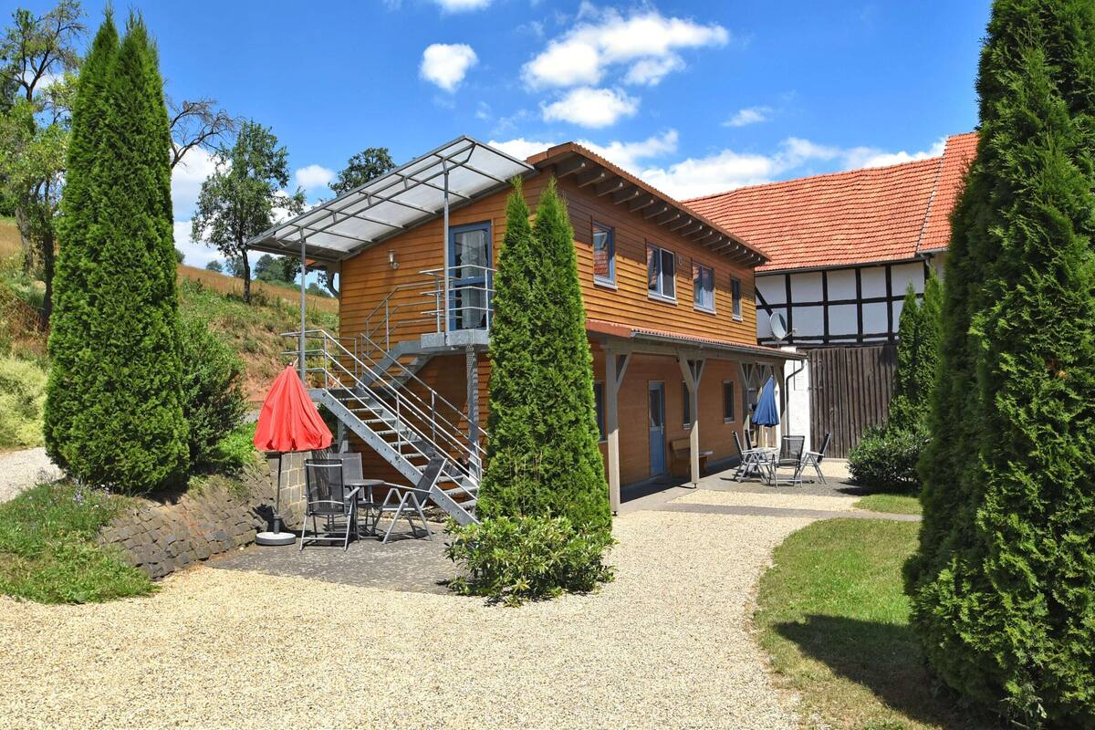 Una casa a due piani in legno con tetto in tegole rosse, un’area salotto esterna coperta e un vialetto in ghiaia.