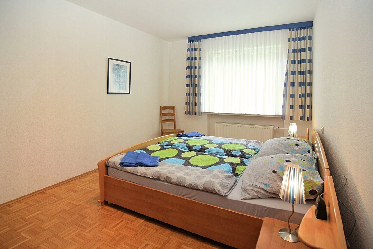 A bedroom with a wooden bed, a nightstand, a chair, a window with curtains, and a framed picture on the wall.