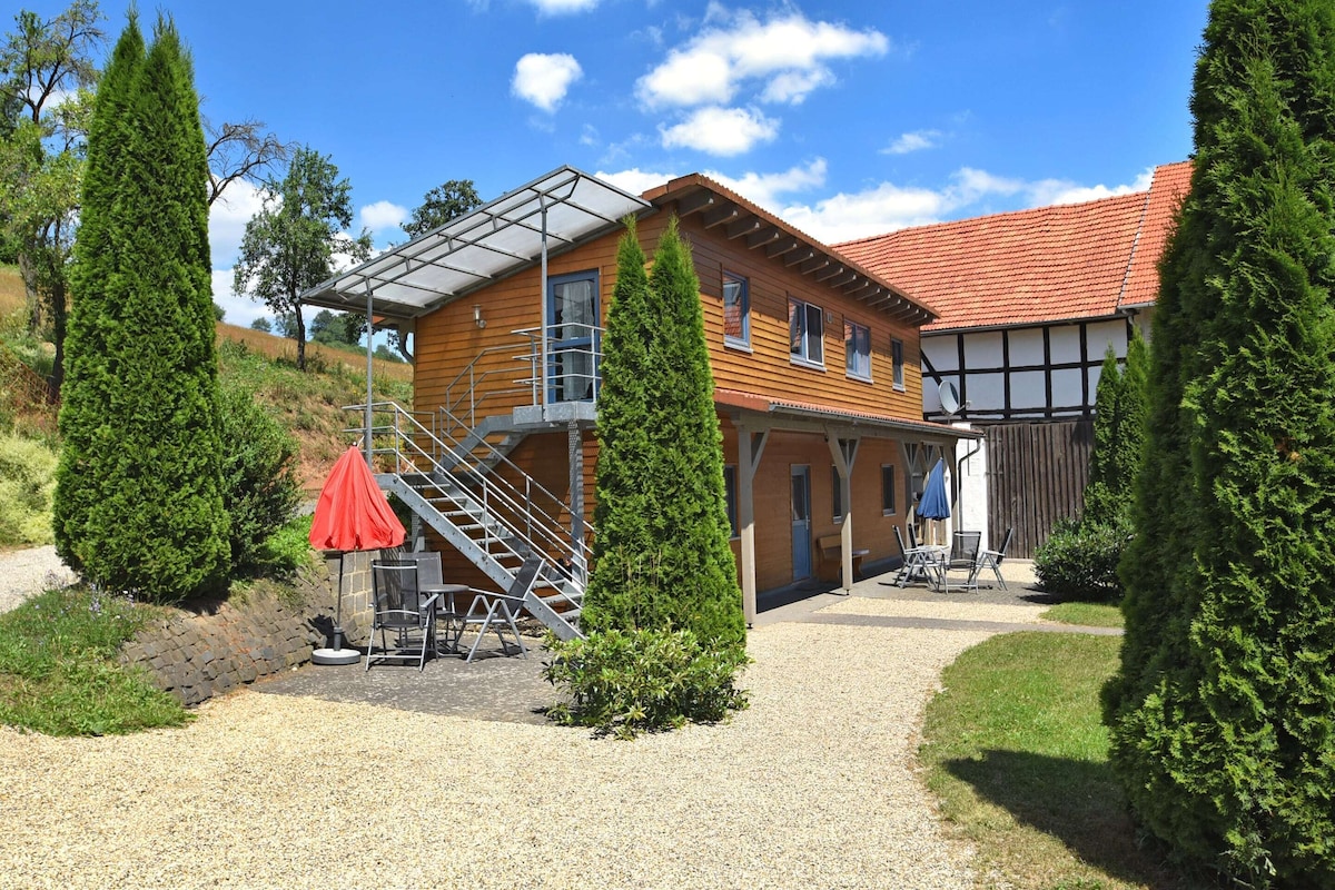 A two-story wooden house with a red-tiled roof, a covered outdoor seating area, and a gravel driveway.