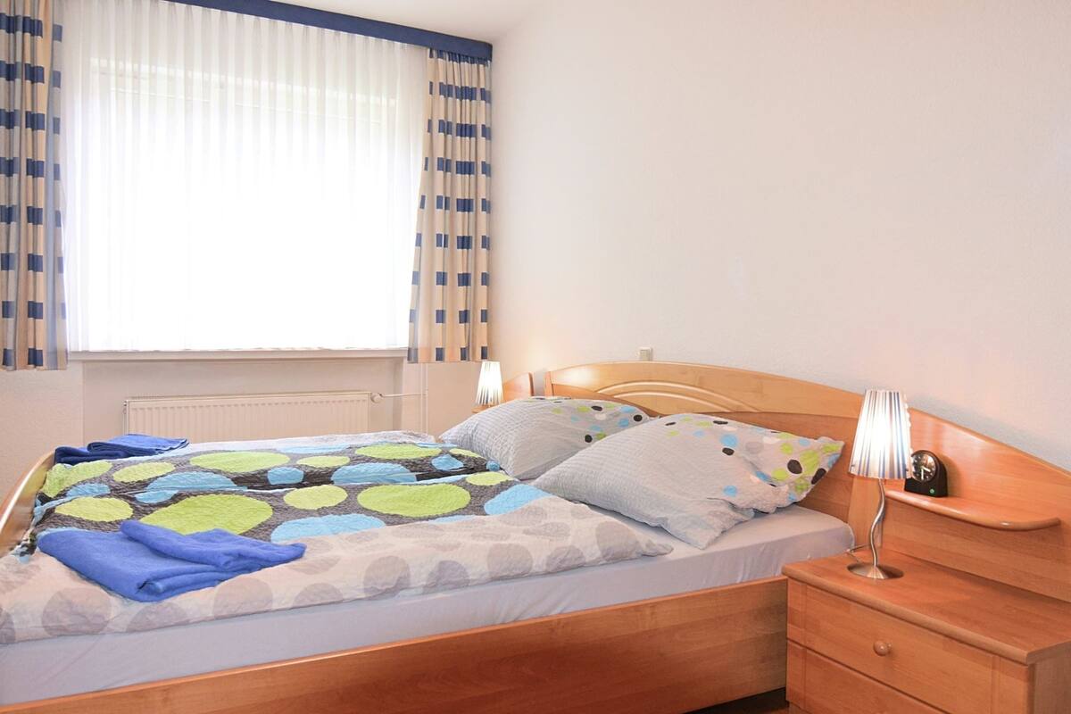 Ein ordentlich bezogenes Bett mit einem gemusterten Überwurf und Kissen, ein Holz-Kopfteil, ein Nachttisch mit Lampe und ein Fenster mit Vorhängen.